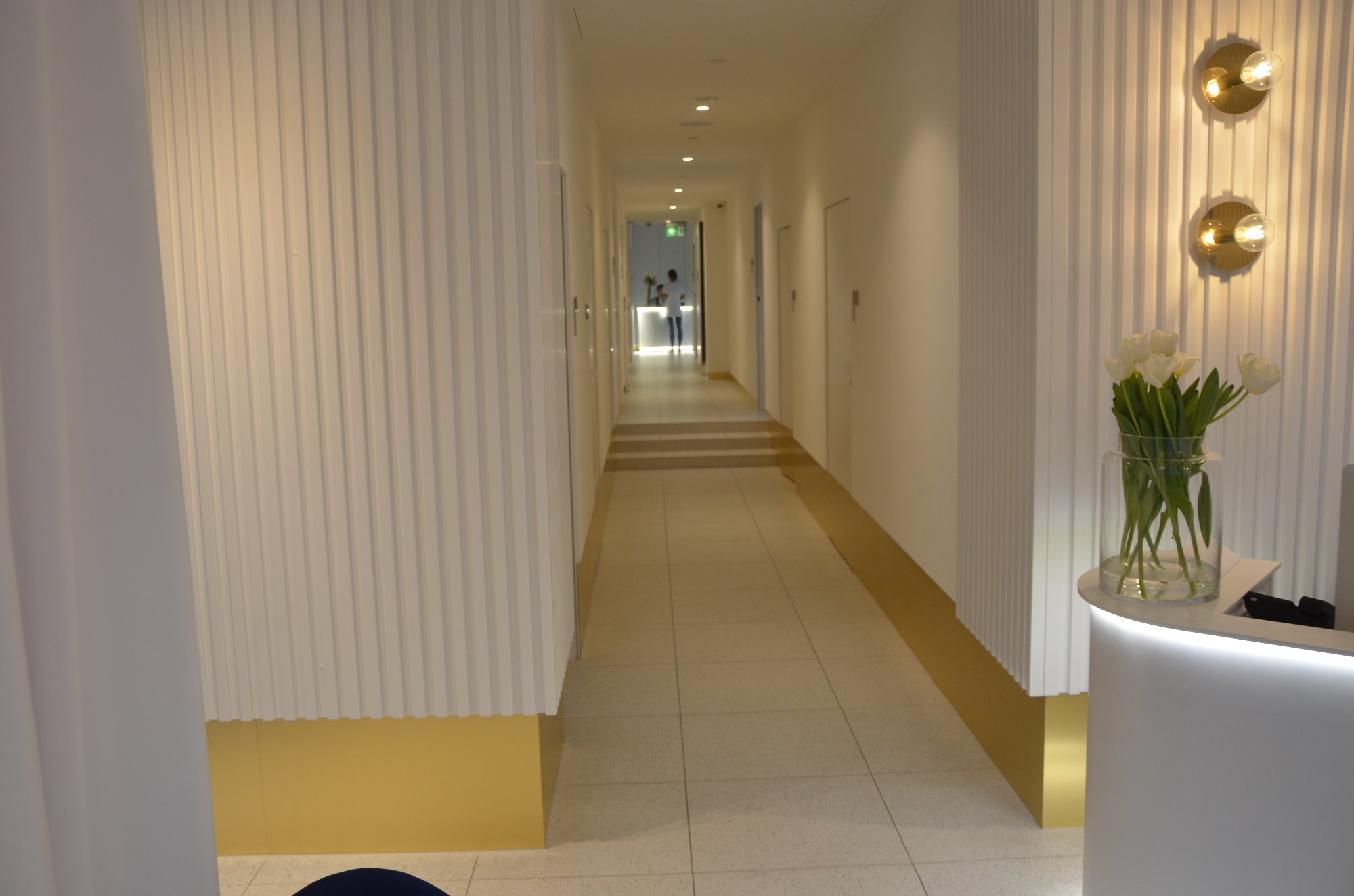 Jasny korytarz biurowy z białymi, żłobkowanymi ścianami, złotymi akcentami u dołu, kwiatami w wazonie na recepcji i widokiem na dalszą część korytarza z postaciami w tle.