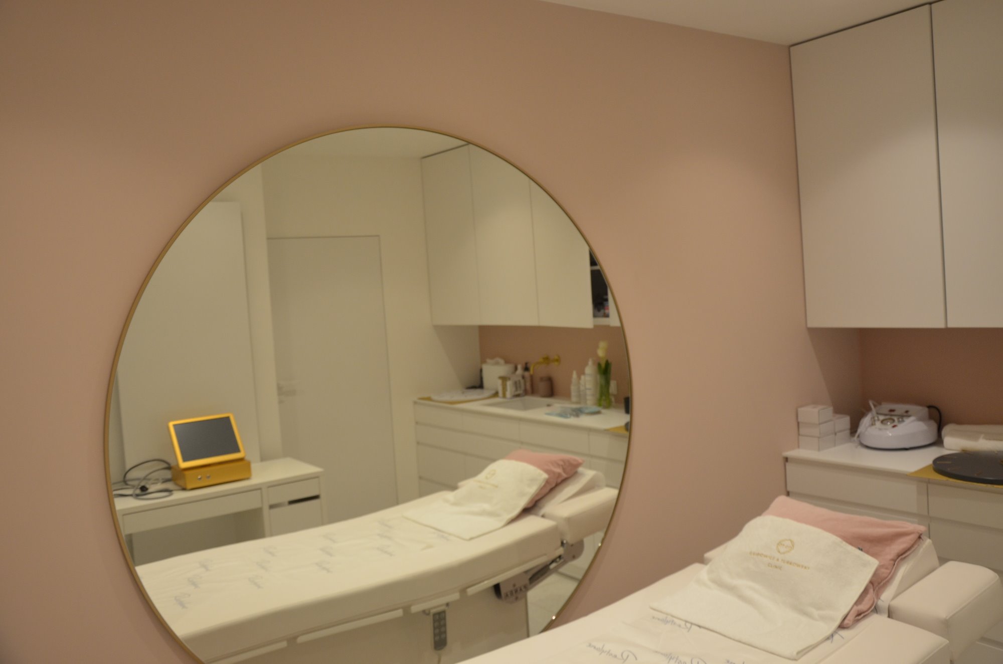 Refleksja w okrągłym lustrze ukazuje wnętrze gabinetu kosmetycznego z dwoma białymi leżankami, jasnoróżowymi ścianami i białymi szafkami. Na jednej z leżanek widoczny ręcznik z logo.