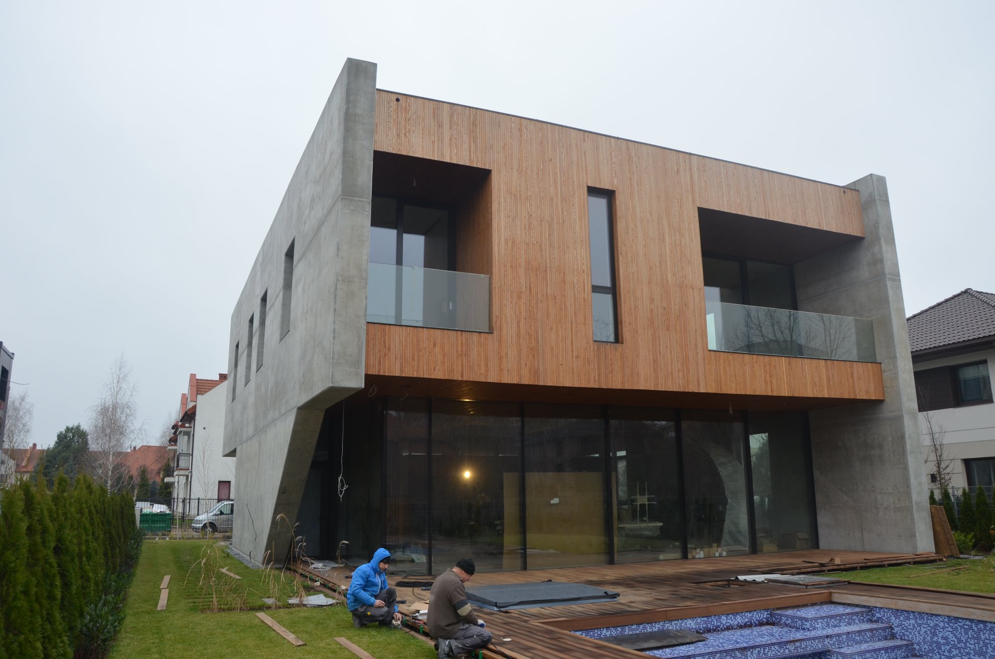 Nowoczesny dom z elementami drewna i betonu, montaż drewnianego tarasu przy basenie, dwóch pracowników w ubraniach roboczych przy pracy, widok z zewnątrz w pochmurny dzień.