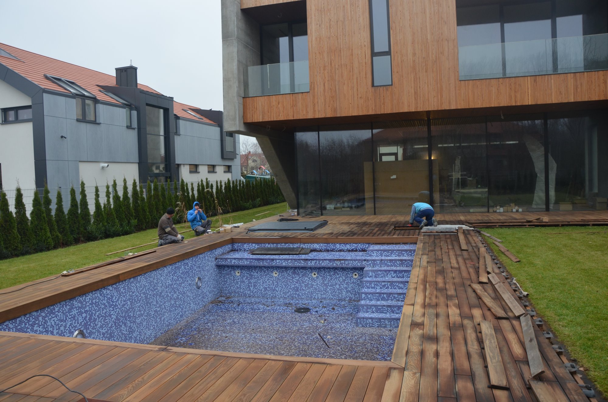 Montaż drewnianego tarasu wokół basenu z niebieską mozaiką w nowoczesnym domu z drewnianą elewacją; dwóch pracowników instaluje deski, trzeci odpoczywa w tle.