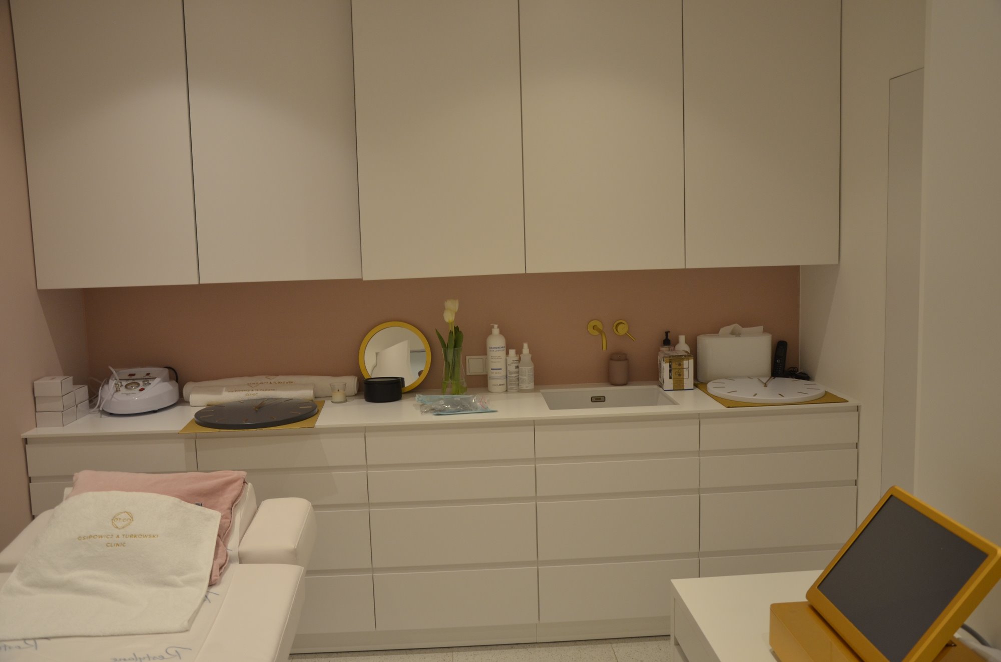 Jasne wnętrze gabinetu kosmetycznego z białymi szafkami, różowym akcentem na ścianie i złotymi detalami, takimi jak lustro i armatura, z leżanką kosmetyczną z ręcznikiem z logo i sprzętem...