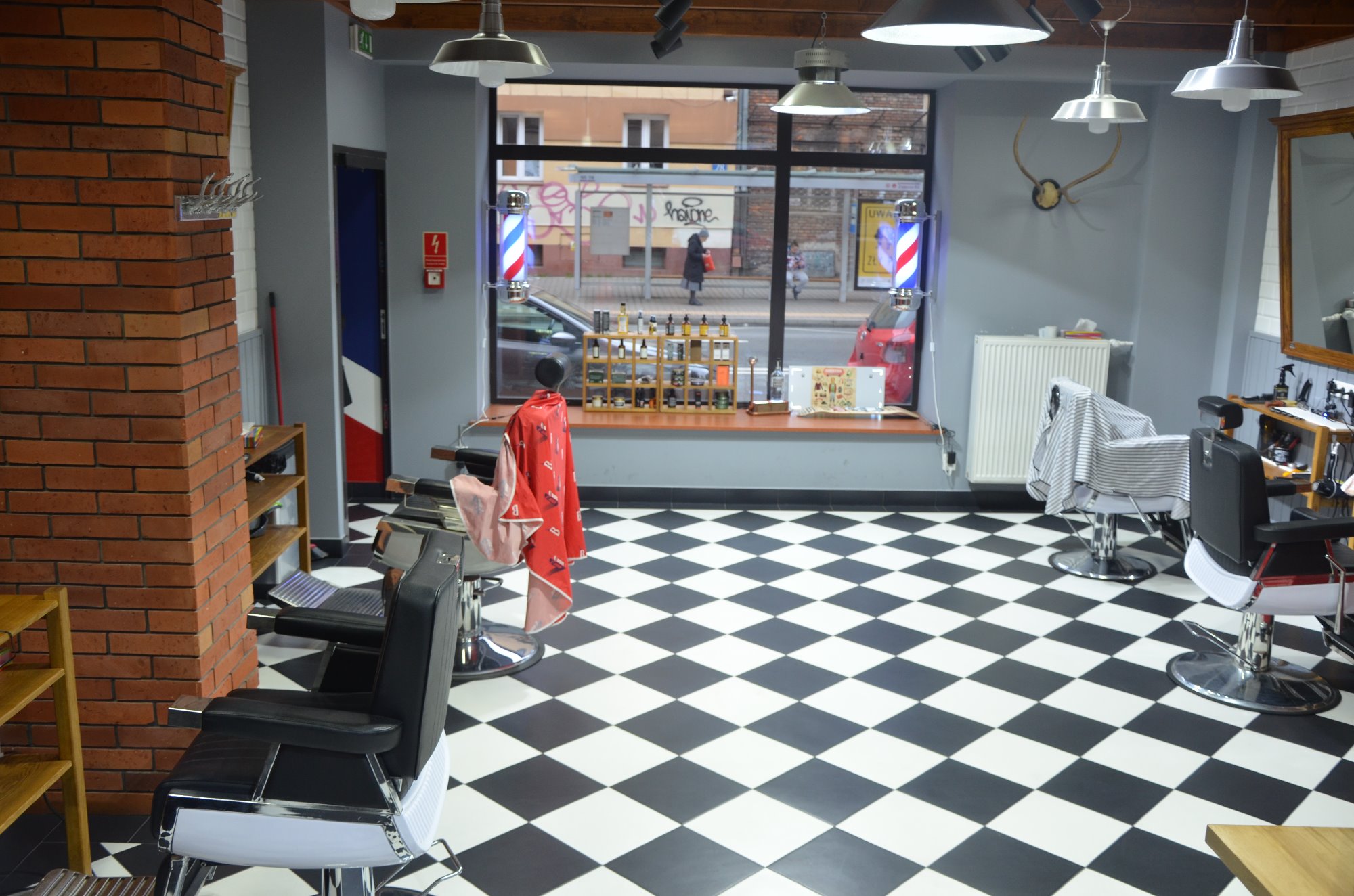 Wnętrze zakładu fryzjerskiego z czarno-białą szachownicą na podłodze, widoczne fotele fryzjerskie, dekoracje ścienne w postaci poroża oraz okno z widokiem na ulicę z charakterystycznymi słupkami...