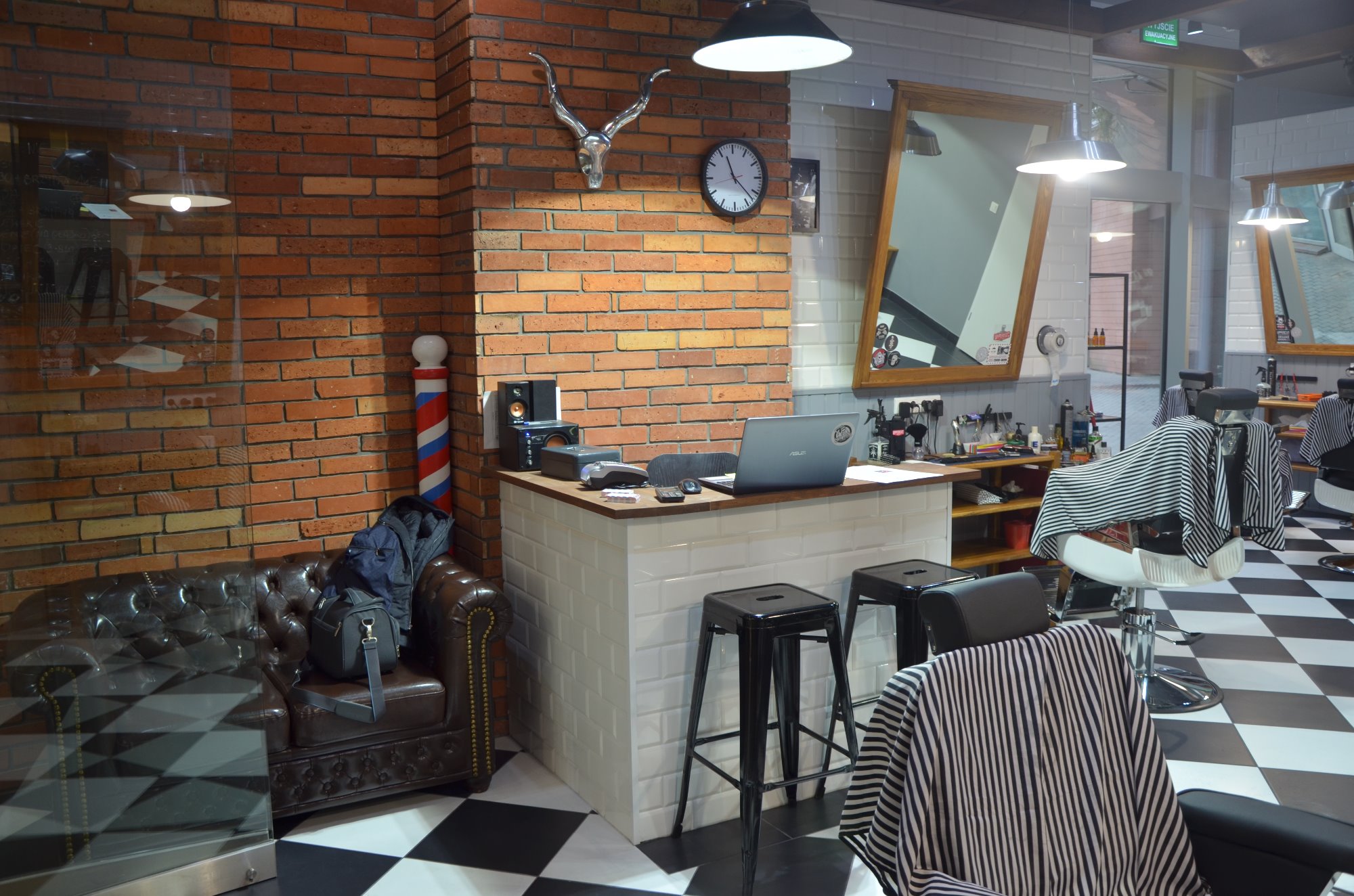 Wnętrze nowoczesnego salonu barberskiego z recepcją wykończoną białymi płytkami, brązową cegłą na ścianie, skórzaną sofą i czarno-białą szachownicą na podłodze, widoczne stanowiska fryzjerskie...