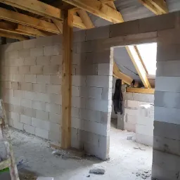 Wnętrze budynku w trakcie murowania ścian z betonu komórkowego, widoczne drewniane elementy konstrukcyjne dachu i prowizoryczna drabina.