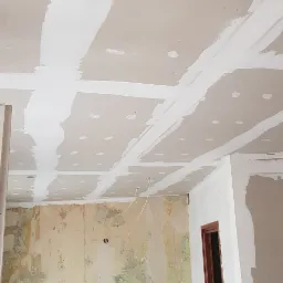 Sufit z płyt kartonowo-gipsowych w trakcie szpachlowania, widoczne łączenia płyt i miejsca po wkrętach, ściana z fragmentami starej farby i nowymi płytkami, bałagan na blacie kuchennym.