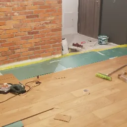 Montaż paneli podłogowych w trakcie realizacji, widoczna podkładka, narzędzia, szczotka do zamiatania, fragment ściany z cegły i drzwi wejściowe.