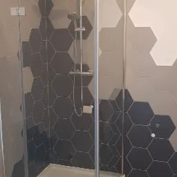 Kabina prysznicowa z deszczownicą, na ścianie heksagonalne płytki w odcieniach szarości, podłoga wyłożona panelami imitującymi drewno.