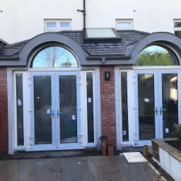 Nowo zainstalowane białe drzwi balkonowe z półokrągłym oknem nad każdym skrzydłem, osadzone w murze z cegły i granitu, widoczne ślady po montażu, w tle biała ściana budynku z oknami i fragment...