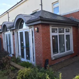 Nowa ceglana dobudówka z białymi oknami i szarym dachem z dachówki, widok z zewnątrz, z elementami wykończeniowymi w trakcie montażu i folią ochronną na szybach.