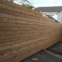 Nowoczesne ogrodzenie z desek ułożonych poziomo, z widocznym fragmentem domu w tle i ziemią u podstawy, świeżo po montażu.