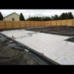 Przygotowany teren pod wylewkę fundamentów, widoczne szalunki, folia izolacyjna i drewniane płyty, w tle ogrodzenie posesji.