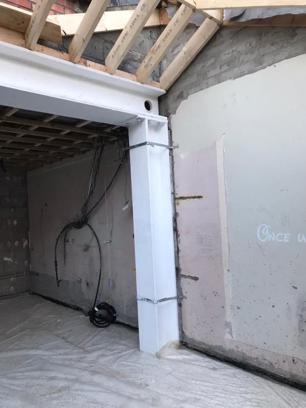 Wnętrze w trakcie prac budowlanych: stalowa belka nośna, wsparta na białej, obudowanej kolumnie, widoczne instalacje elektryczne, zabezpieczona folią podłoga i surowy stan ścian.