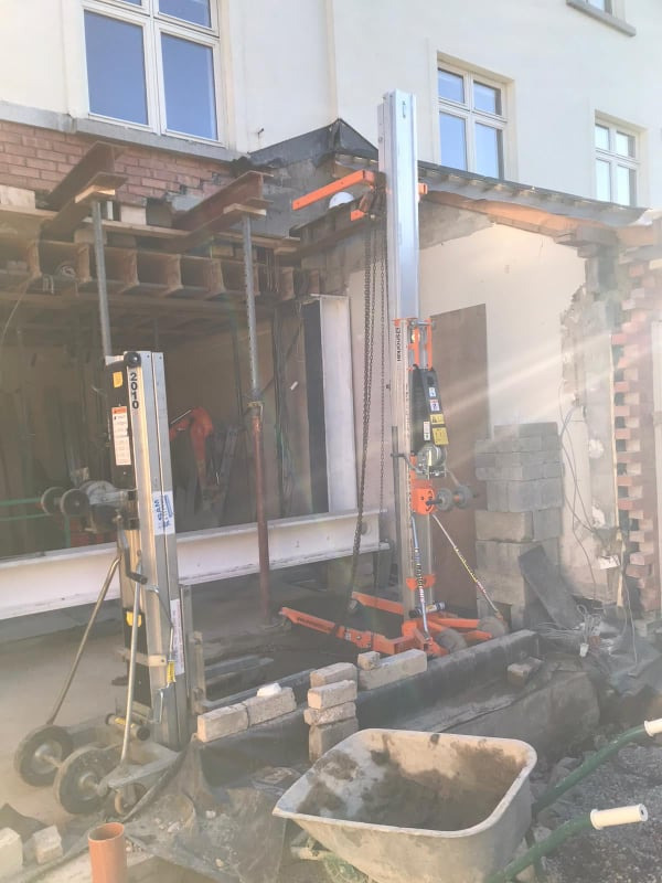 Montaż stalowych belek wzmacniających nadproże okienne z użyciem podnośników teleskopowych; widoczna ceglana elewacja budynku w trakcie przebudowy i wózek budowlany z zaprawą.