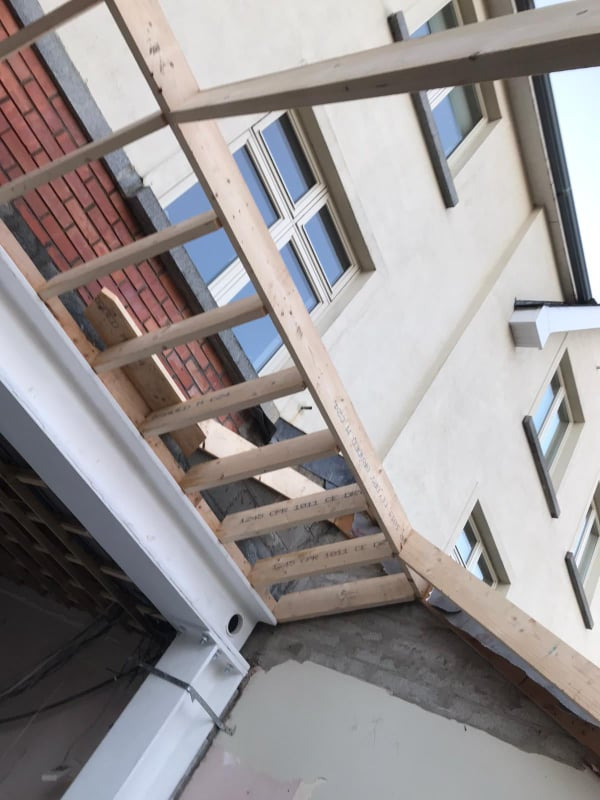 Szkielet konstrukcji drewnianej, prawdopodobnie zadaszenia, widoczny od dołu, z fragmentem ściany budynku z oknami w tle. Widoczna cegła i surowy tynk.