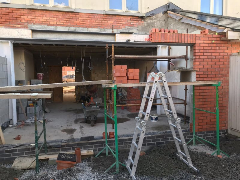 Budowa domu w trakcie wznoszenia ścian z czerwonej cegły, widoczne rusztowania, drabina i materiały budowlane, stan surowy otwarty.