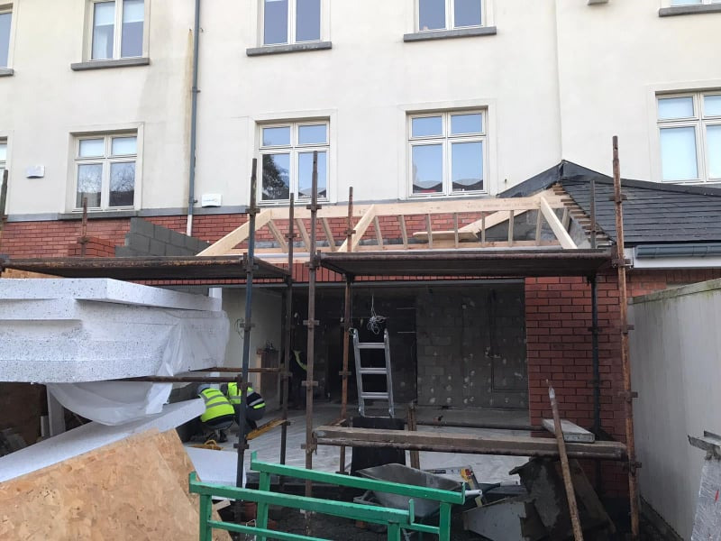 Budowa nowej części budynku z czerwonej cegły, widoczne rusztowania, drewniana konstrukcja dachu, izolacja termiczna i pracownicy w kamizelkach odblaskowych.