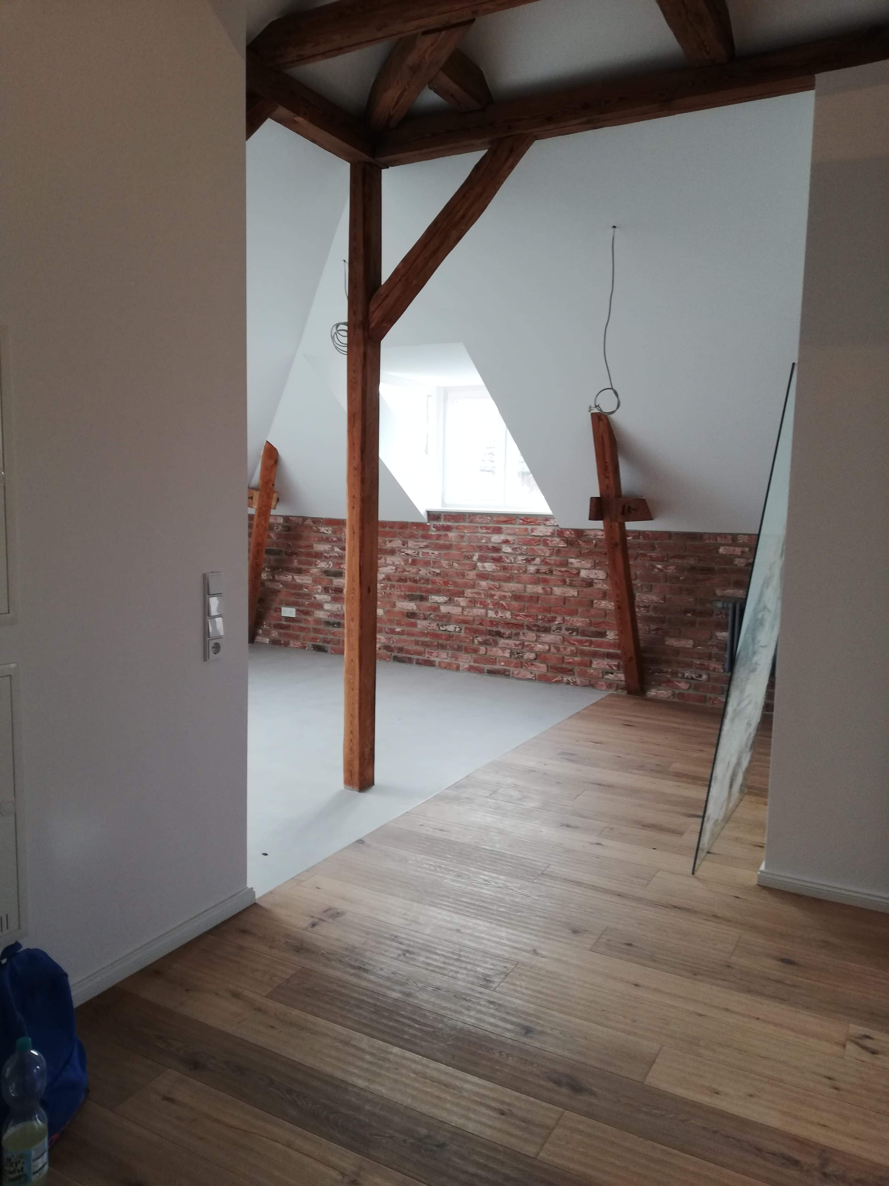 Poddasze po remoncie: drewniane belki stropowe, ściana z cegły, jasna podłoga drewniana i wylewka samopoziomująca, widok z korytarza.