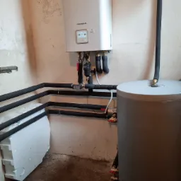 Biały piec grzewczy LG Therma V z wyświetlaczem, podłączony do rur w izolacji termicznej i szarego zasobnika wody w surowym wnętrzu kotłowni.