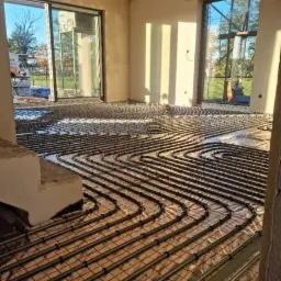 Rozłożone rury ogrzewania podłogowego na folii izolacyjnej z siatką w surowym wnętrzu domu w trakcie budowy, widok z niskiej perspektywy.