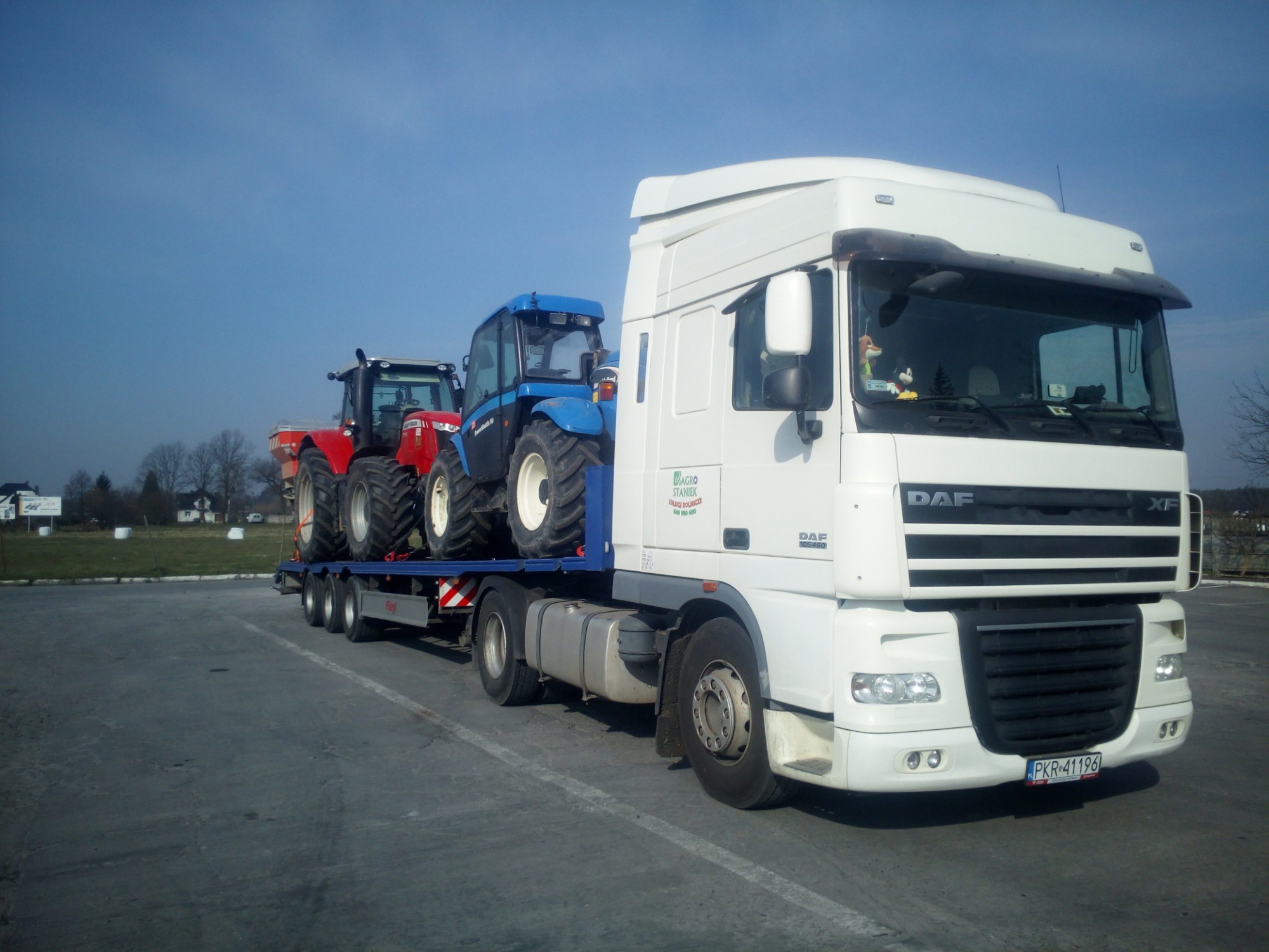 Ciężarówka DAF z naczepą niskopodwoziową przewożąca dwa traktory rolnicze: czerwony i niebieski, zaparkowana na placu.