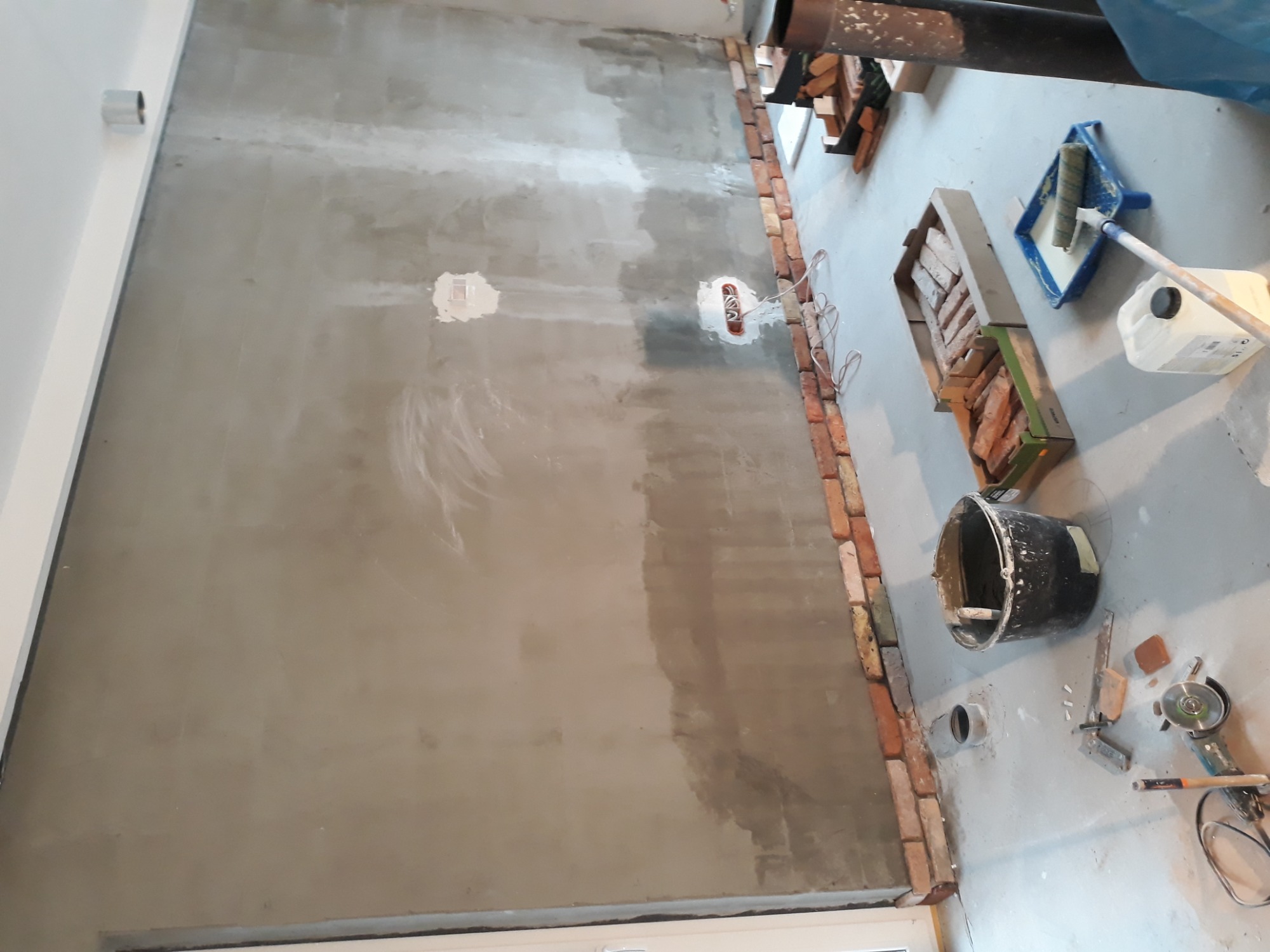 Ściana w trakcie murowania z widoczną warstwą zaprawy, częściowo obłożona czerwoną cegłą, z widocznymi puszkami elektrycznymi, narzędziami oraz pojemnikami z cegłami i farbą.