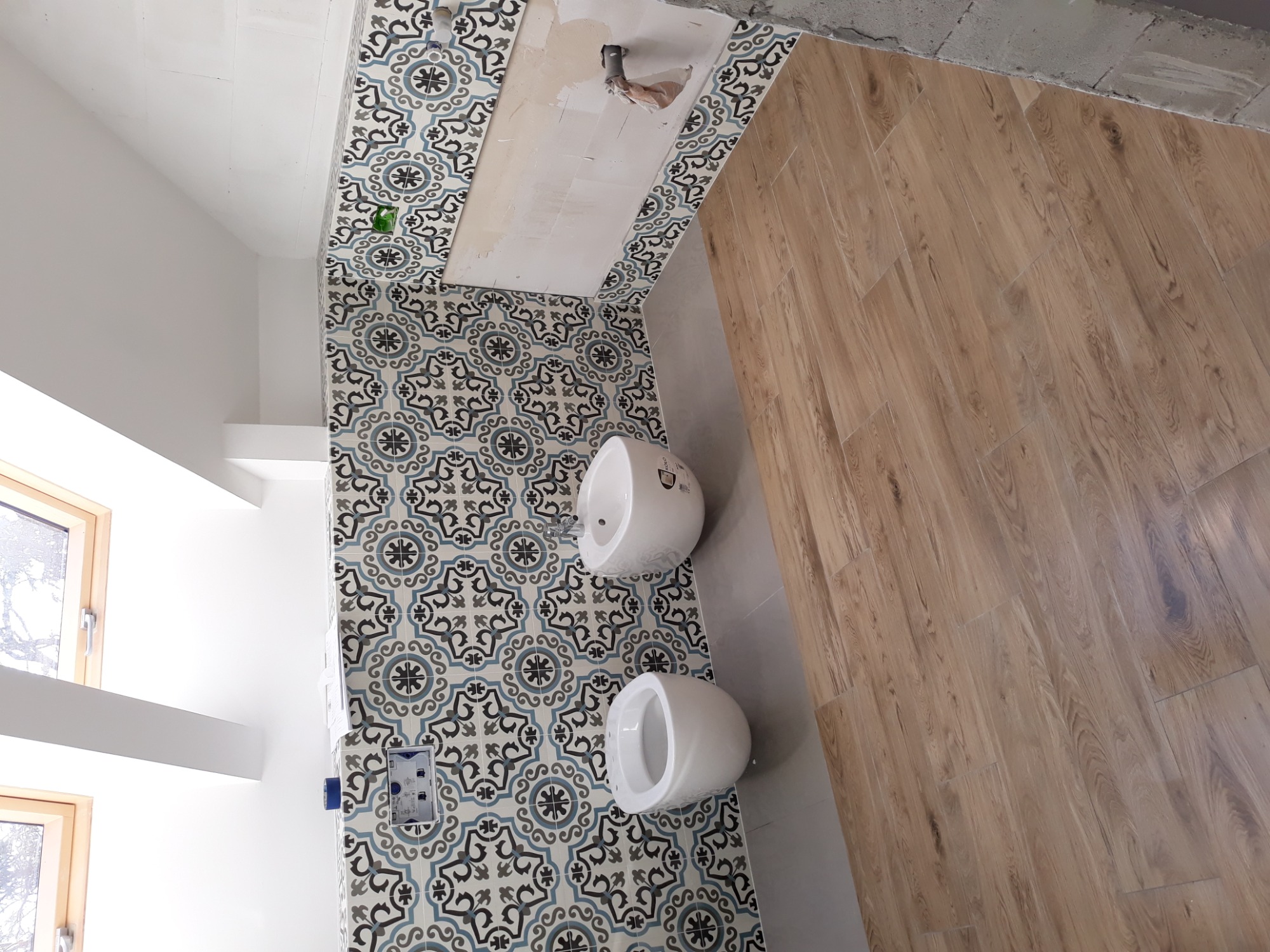 Łazienka w trakcie wykańczania: ułożone płytki podłogowe imitujące drewno, ściany z dekoracyjnymi płytkami w geometryczne wzory, zamontowane białe bidety, widoczne puszki elektryczne...