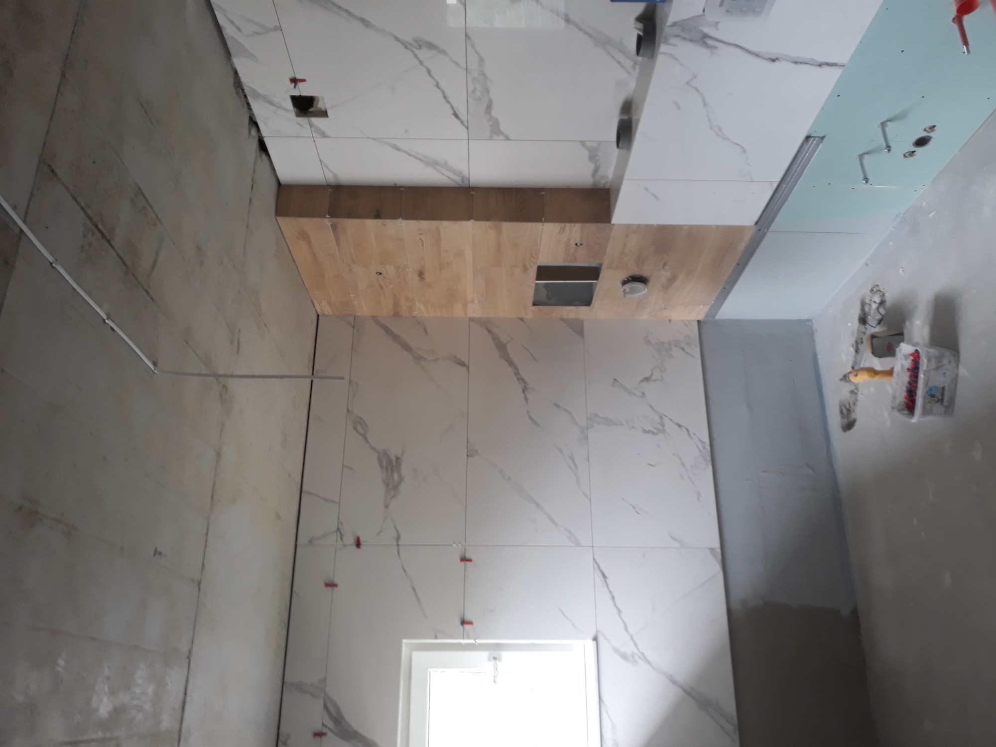 Łazienka w trakcie remontu: białe płytki imitujące marmur, drewniane elementy zabudowy z otworem na oświetlenie, widoczne krzyżyki dystansowe, surowa ściana i narzędzia glazurnicze.