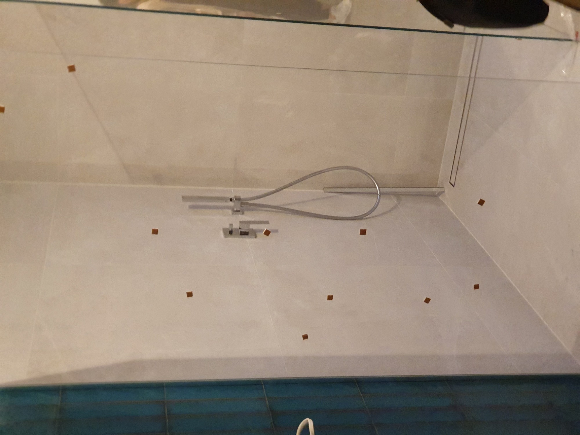 Świeżo wykafelkowana ściana kabiny prysznicowej z zamontowaną baterią i słuchawką, zabezpieczona brązowymi podkładkami dystansowymi, dolna część wykończona niebieską mozaiką.