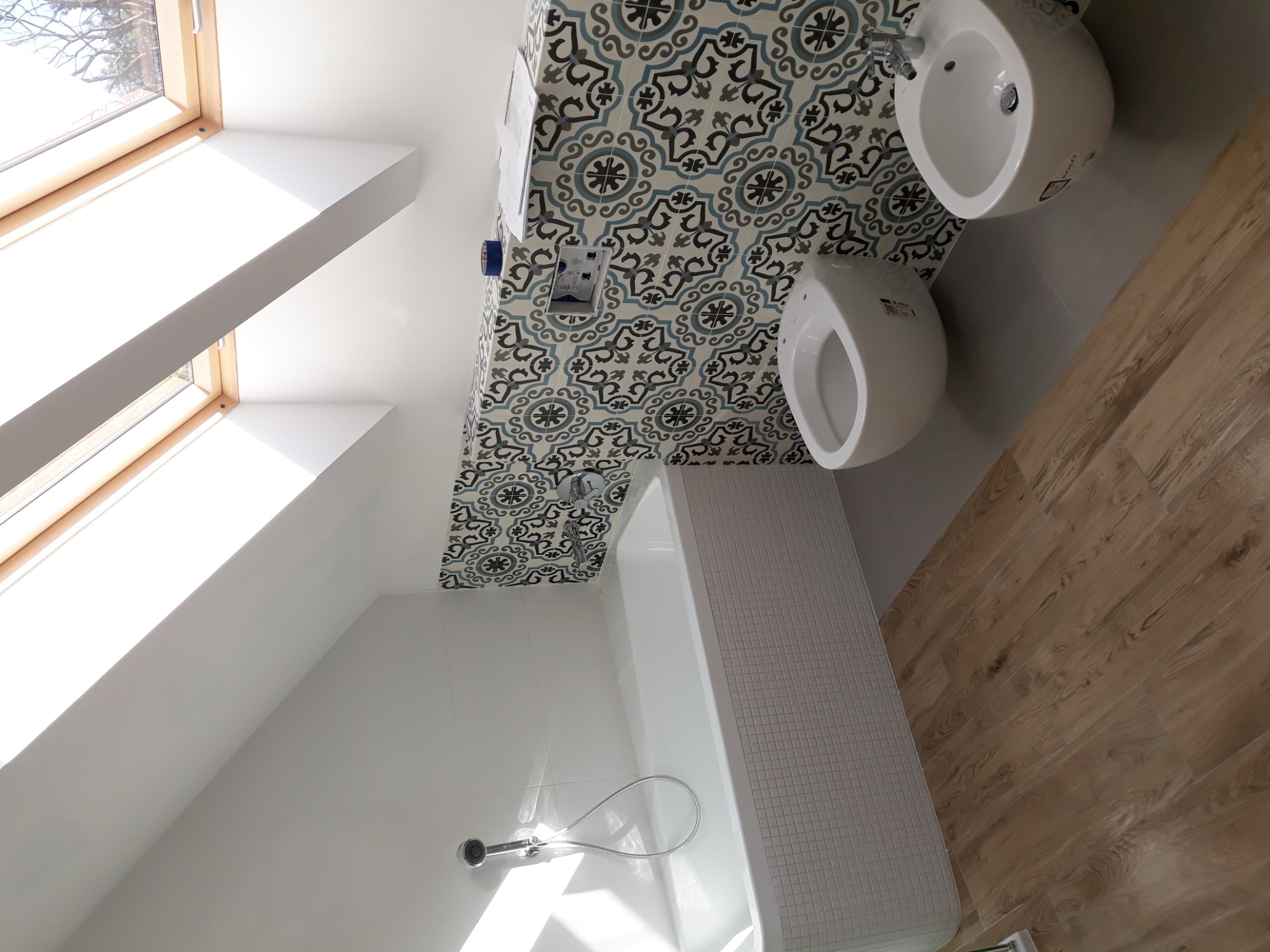 Jasna łazienka na poddaszu z oknem dachowym, białą wanną obłożoną mozaiką, umywalką, bidetem i wzorzystymi płytkami na ścianie.