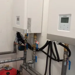 Dwa białe urządzenia Kaisai zamontowane na ścianie, połączone rurami z zaworami i czarnymi przewodami, z czerwonym zbiornikiem wyrównawczym na pierwszym planie.