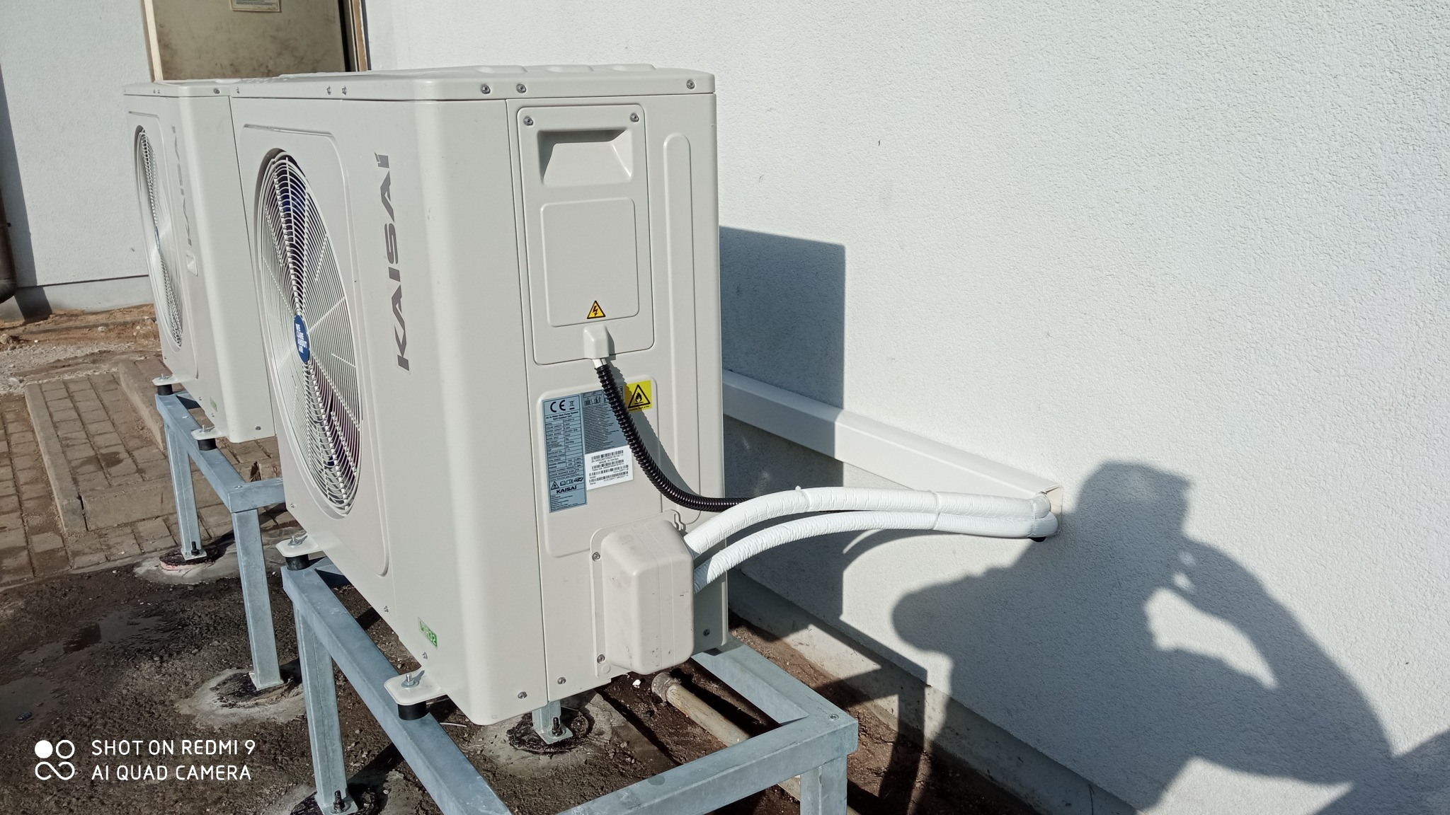 Dwa zewnętrzne moduły klimatyzacji Kaisai zamontowane na metalowych stelażach przy białej ścianie budynku, widoczne rury w izolacji i cień osoby fotografującej.