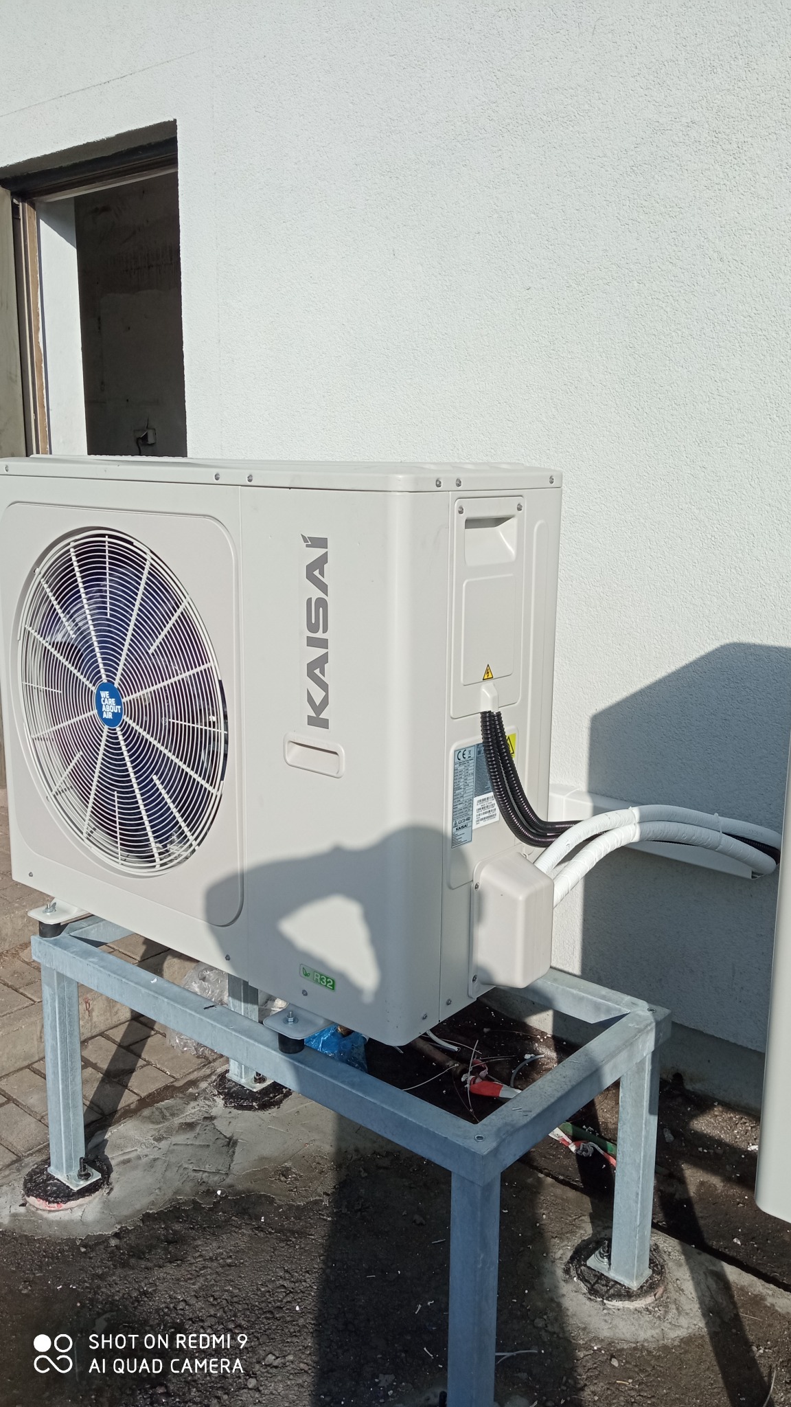 Zewnętrzna jednostka klimatyzacji Kaisai na metalowym stelażu, widoczne przyłącza przewodów w białej izolacji i czarnej karbowanej osłonie, tło stanowi biała ściana budynku z otwartym wjazdem.