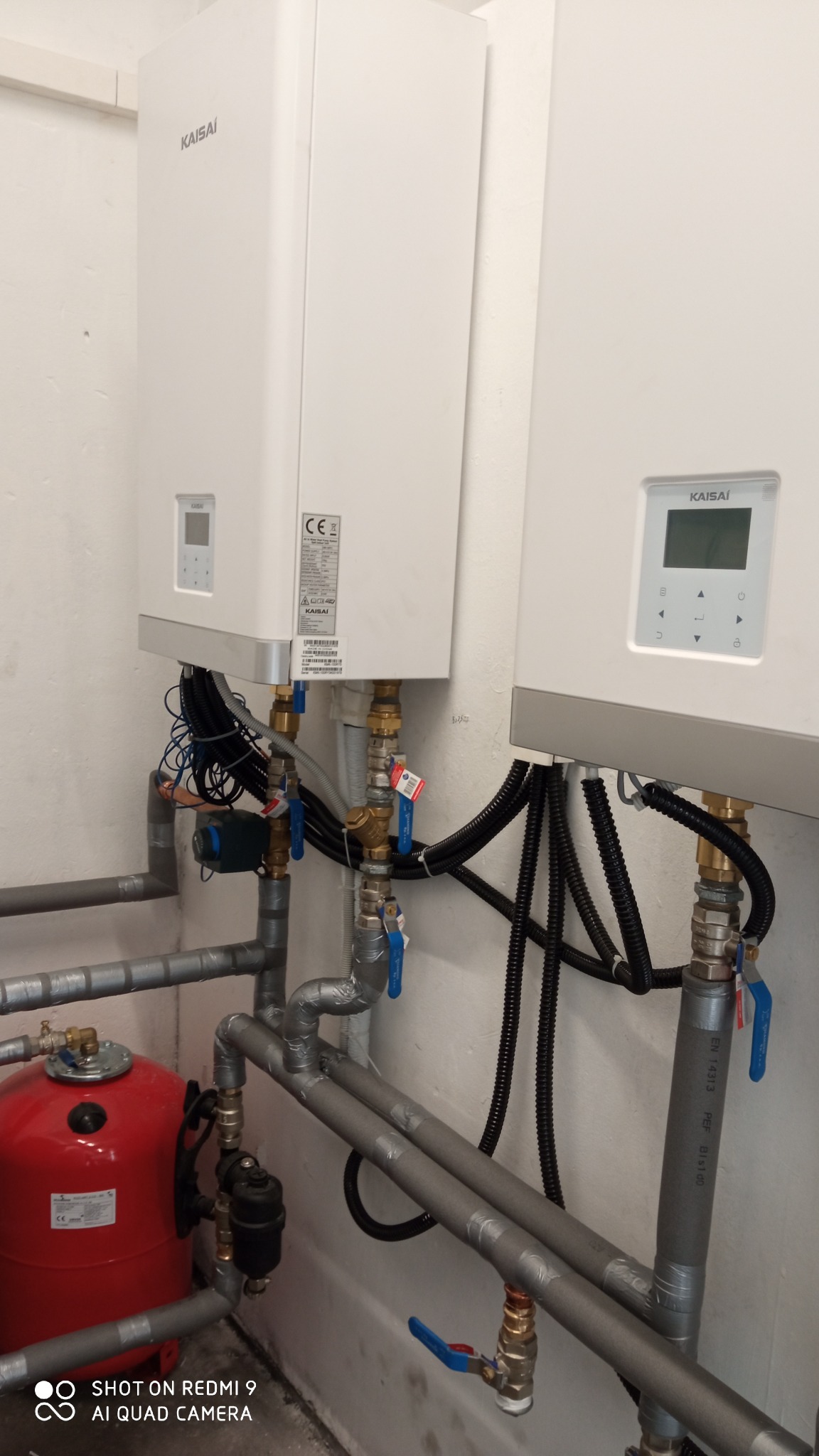 Dwa białe urządzenia Kaisai zamontowane na ścianie, połączone rurami z zaworami i czarnymi przewodami, z czerwonym zbiornikiem wyrównawczym na pierwszym planie.