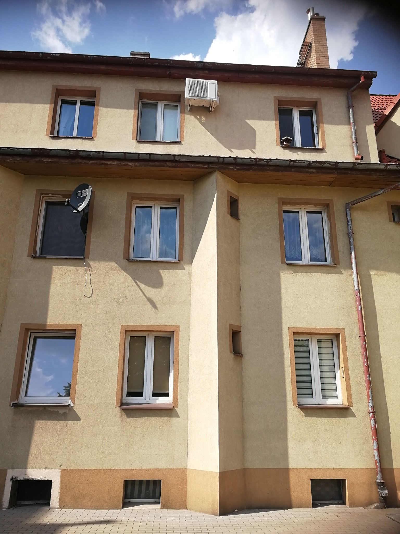 Elewacja budynku z zamontowaną klimatyzacją zewnętrzną, widoczne okna z roletami i satelita na ścianie.