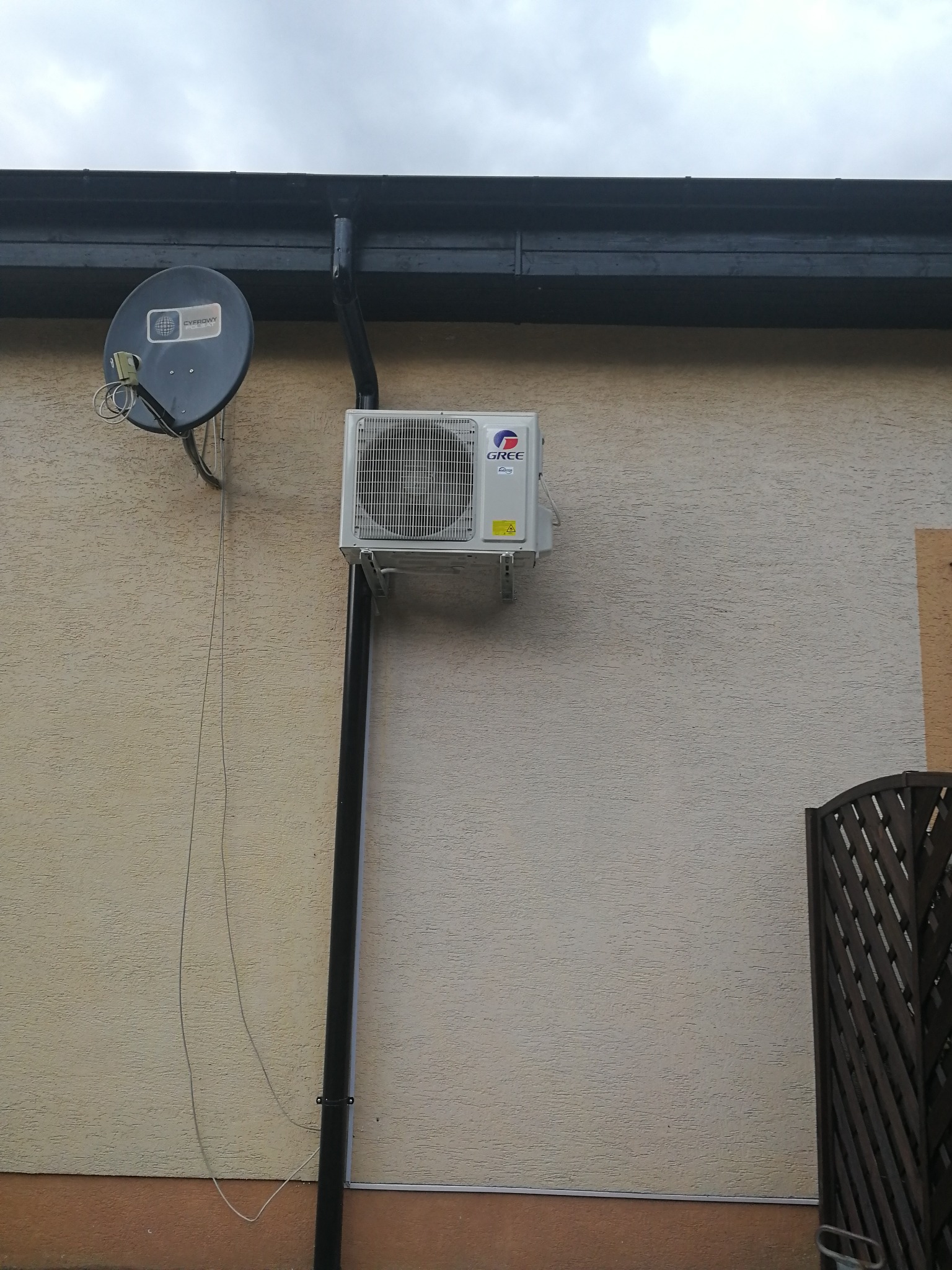 Zewnętrzna jednostka klimatyzacji Gree zamontowana na ścianie budynku obok rynny spustowej i anteny satelitarnej.