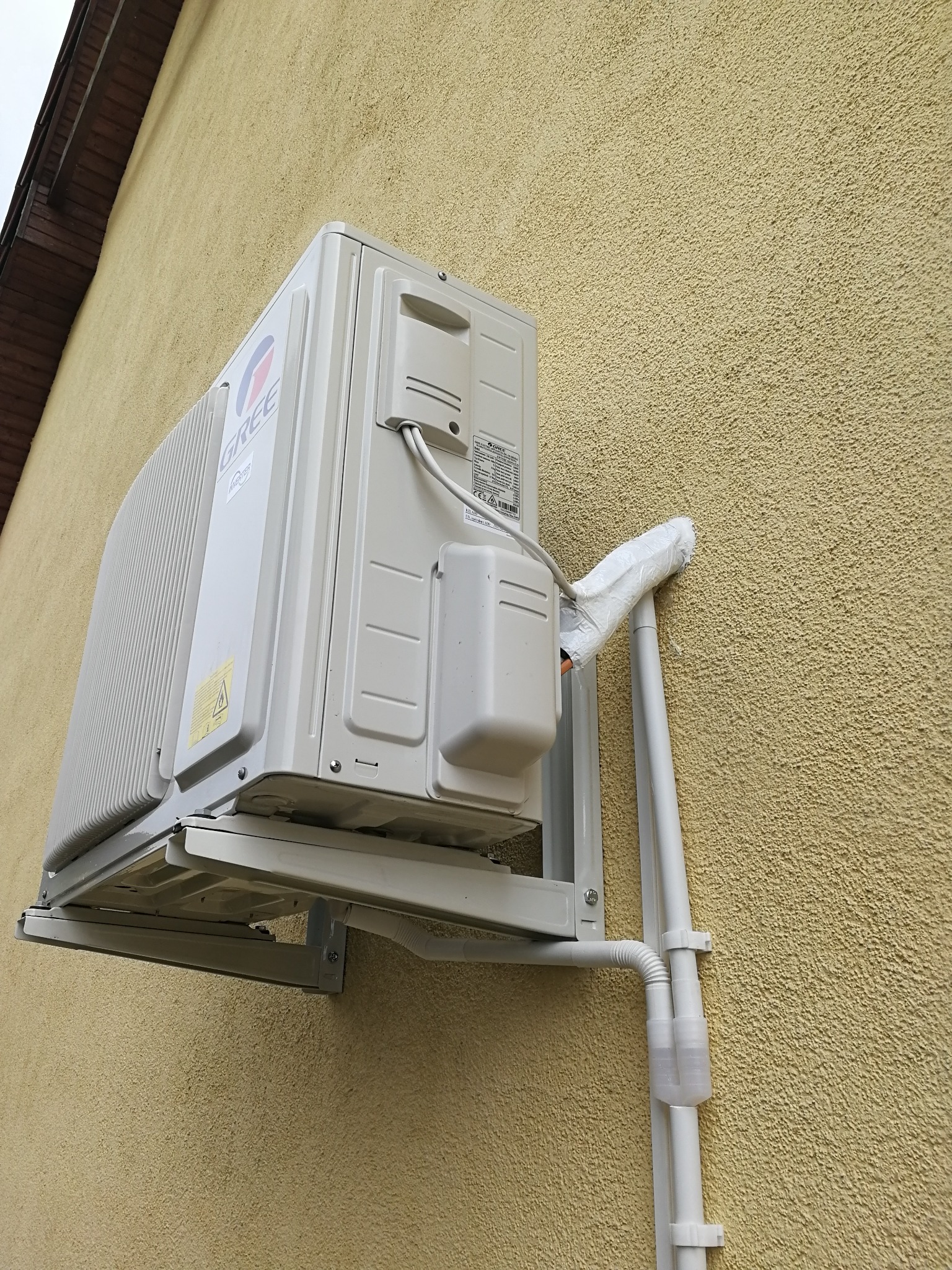 Zewnętrzna jednostka klimatyzacji marki Gree zamontowana na żółtej, chropowatej ścianie budynku, z widocznymi rurami odprowadzającymi skropliny owiniętymi białą taśmą.