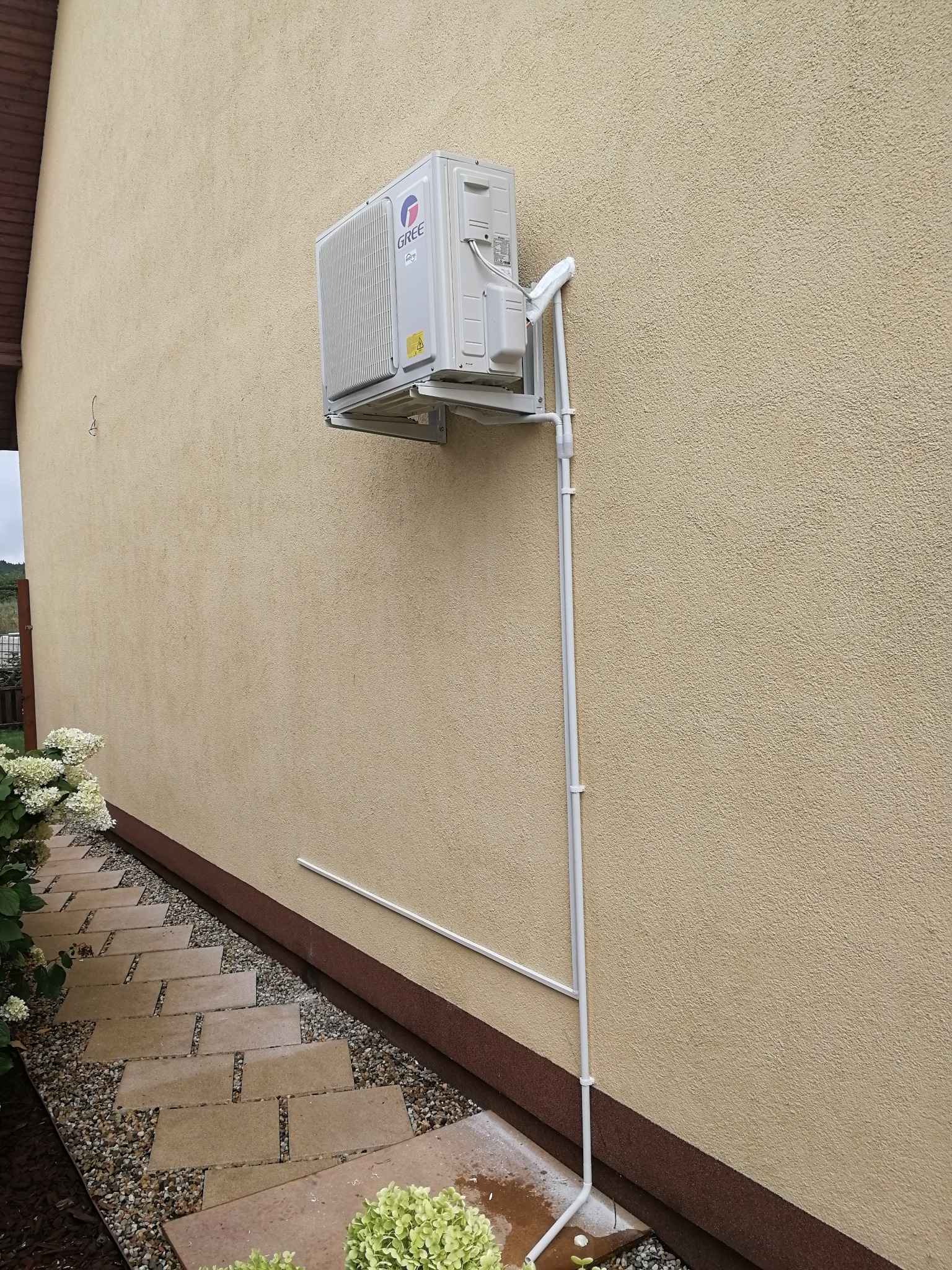 Zewnętrzna jednostka klimatyzacji marki Gree zamontowana na elewacji domu, z widocznymi rurami odprowadzającymi skropliny i chodnikiem z kostki brukowej w tle.