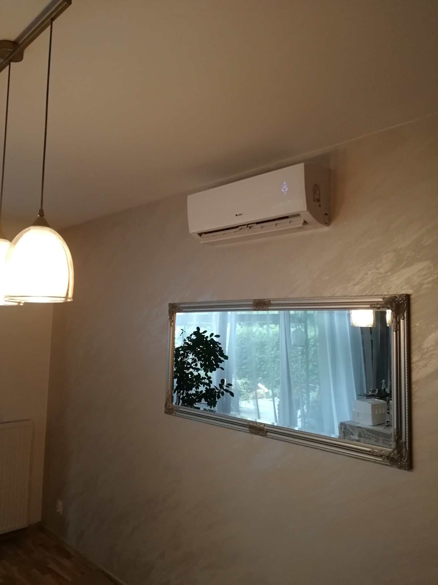Biały klimatyzator zamontowany na ścianie nad ozdobnym lustrem, w tle widoczne lampy wiszące i grzejnik.