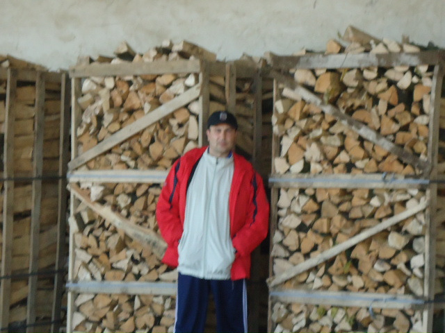 Mężczyzna w czerwonej kurtce stoi przed paletami z pociętym drewnem opałowym w magazynie.