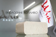 Kostka jasnego brykietu drzewnego na szklanym blacie, w tle flaga z logo firmy i napis 'EKOLOGICZNE PALIWO'.