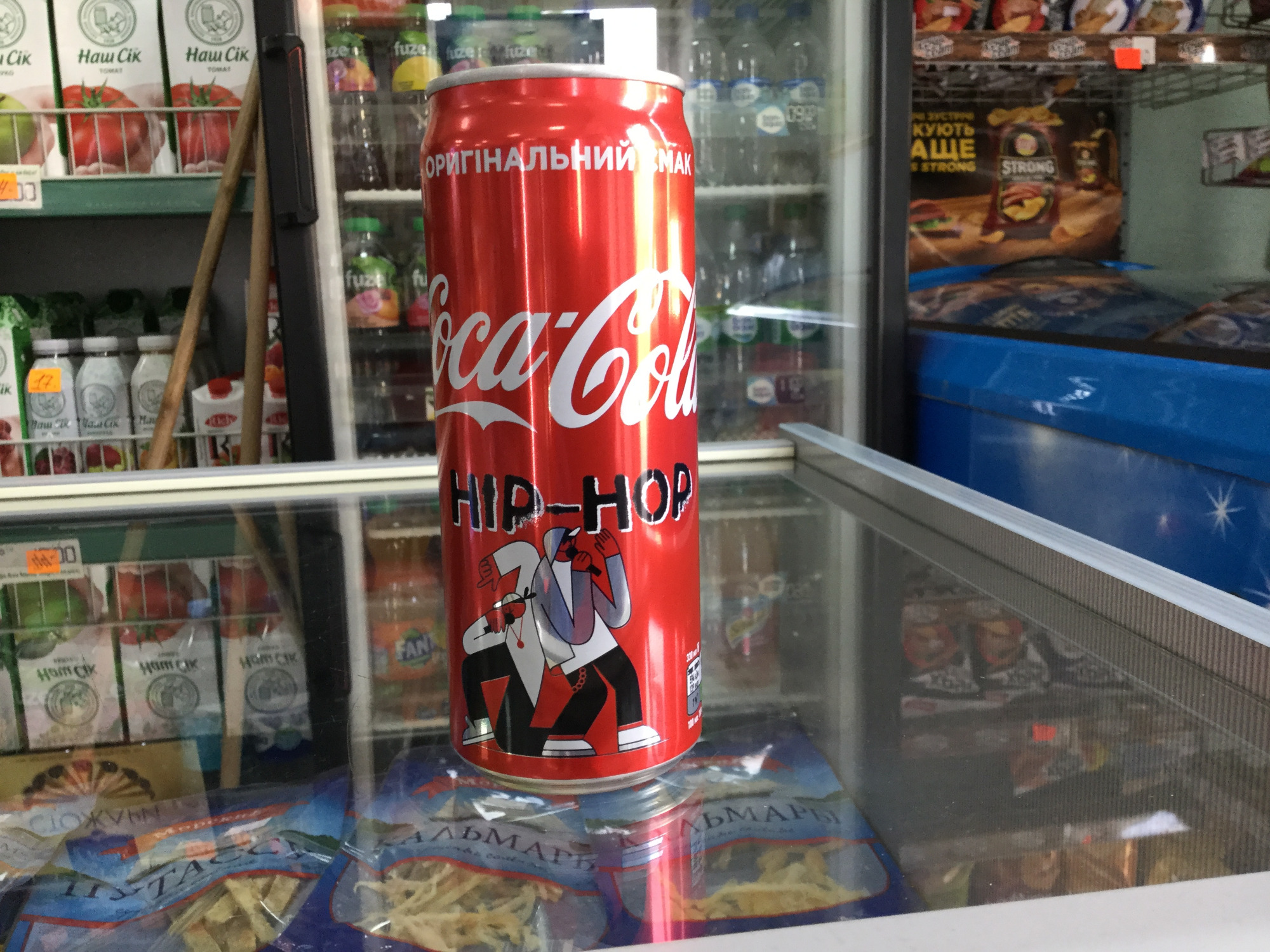 Puszka Coca-Coli z grafiką w stylu hip-hop, umieszczona na szklanej powierzchni lady chłodniczej, widoczne produkty spożywcze w tle.