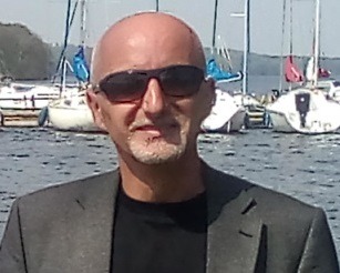 Portret mężczyzny w okularach przeciwsłonecznych i marynarce na tle przystani z jachtami, lekko prześwietlone zdjęcie.