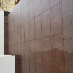 Brązowe płytki ceramiczne ułożone na zewnątrz budynku, widoczne krzyżyki dystansowe i piasek przy krawędziach, biała ściana z widoczną fakturą obok.