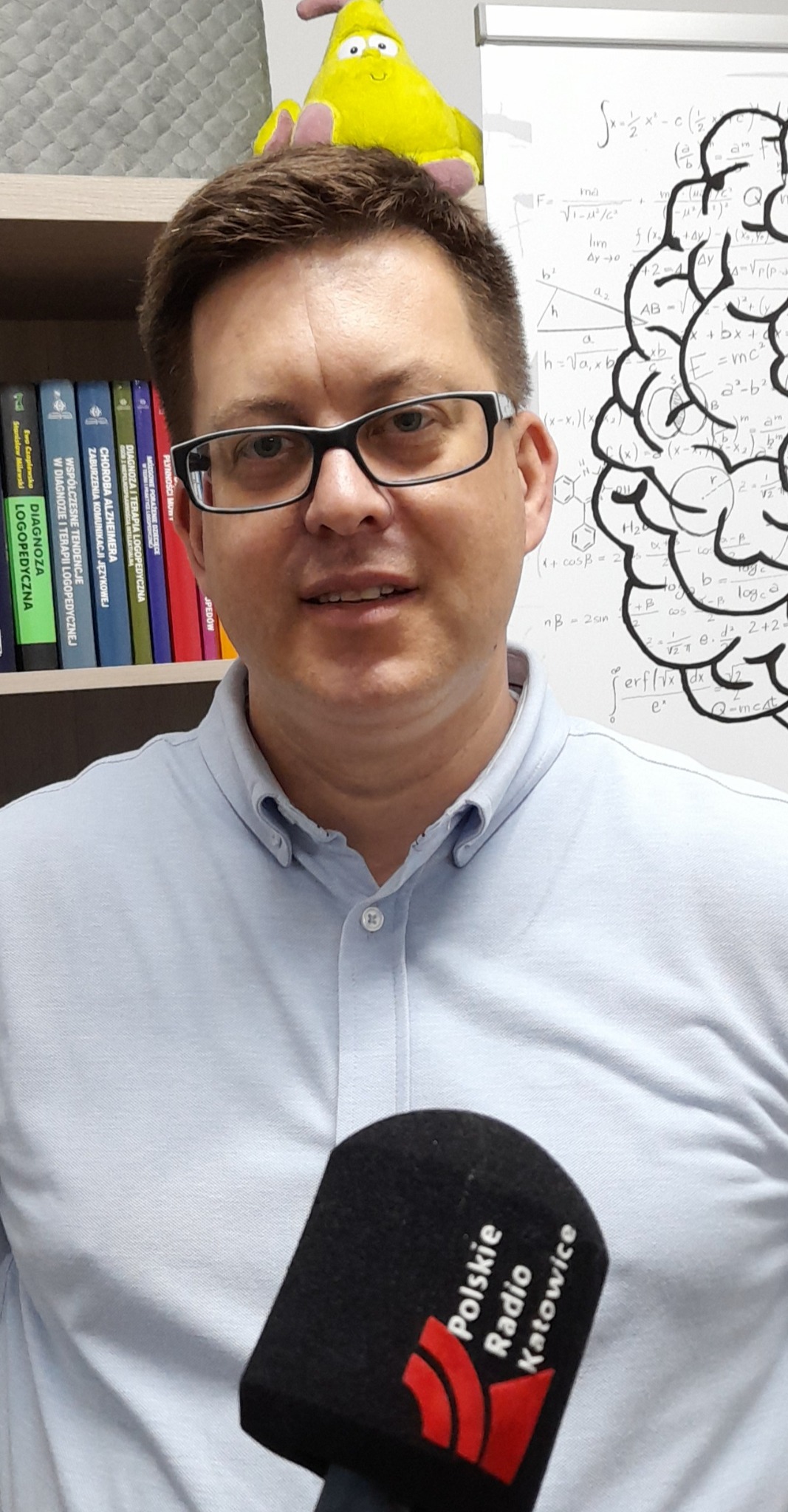 Mężczyzna w okularach, z mikrofonem Polskiego Radia Katowice, na tle półki z książkami o logopedii i tablicy z rysunkiem mózgu i wzorami matematycznymi.