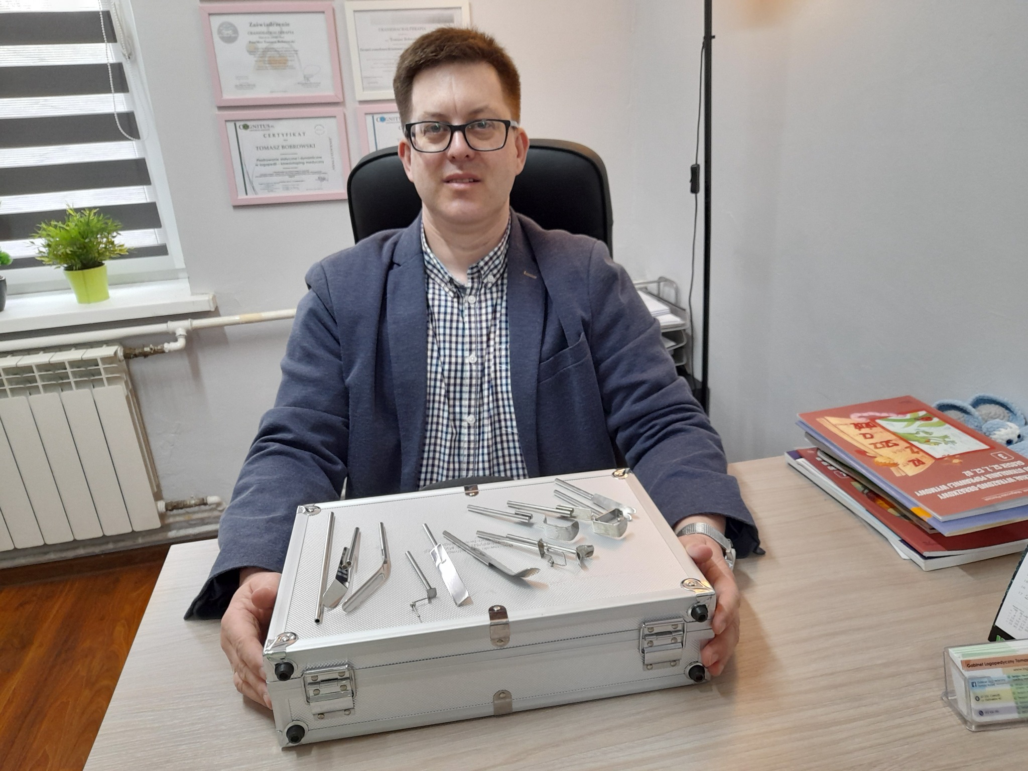 Mężczyzna w okularach i marynarce siedzi za biurkiem, na którym leży srebrna walizka z zestawem narzędzi logopedycznych, w tle certyfikaty na ścianie i roślina doniczkowa na parapecie.