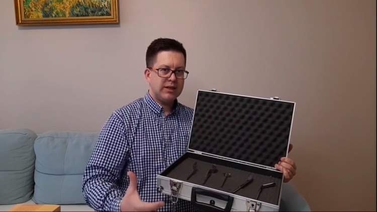 Mężczyzna w okularach prezentuje srebrną walizkę z zestawem narzędzi diagnostycznych, siedząc na jasnoniebieskiej kanapie z obrazem w złotej ramie w tle.