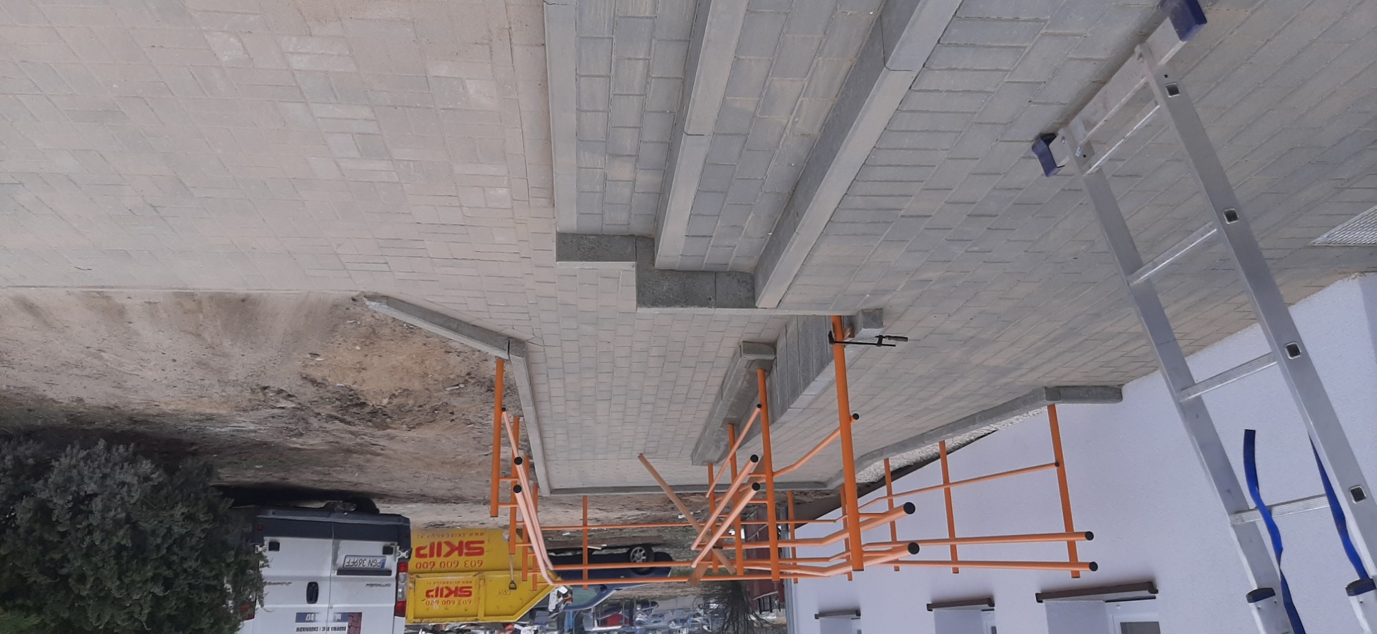 Trwa układanie jasnoszarej kostki brukowej na schodach wejściowych do budynku; widoczne rusztowanie z pomarańczowych rurek oraz drabina oparta o ścianę budynku.