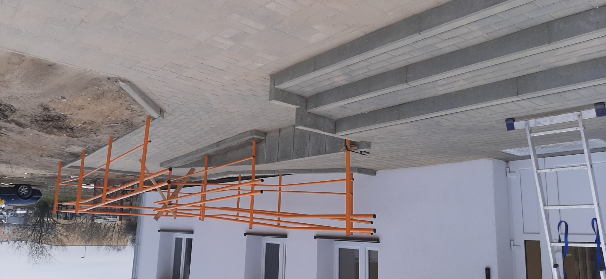 Ułożona szara kostka brukowa tworząca schody i chodnik, z widocznym rusztowaniem pomalowanym na pomarańczowo i aluminiową drabiną opartą o białą ścianę budynku.