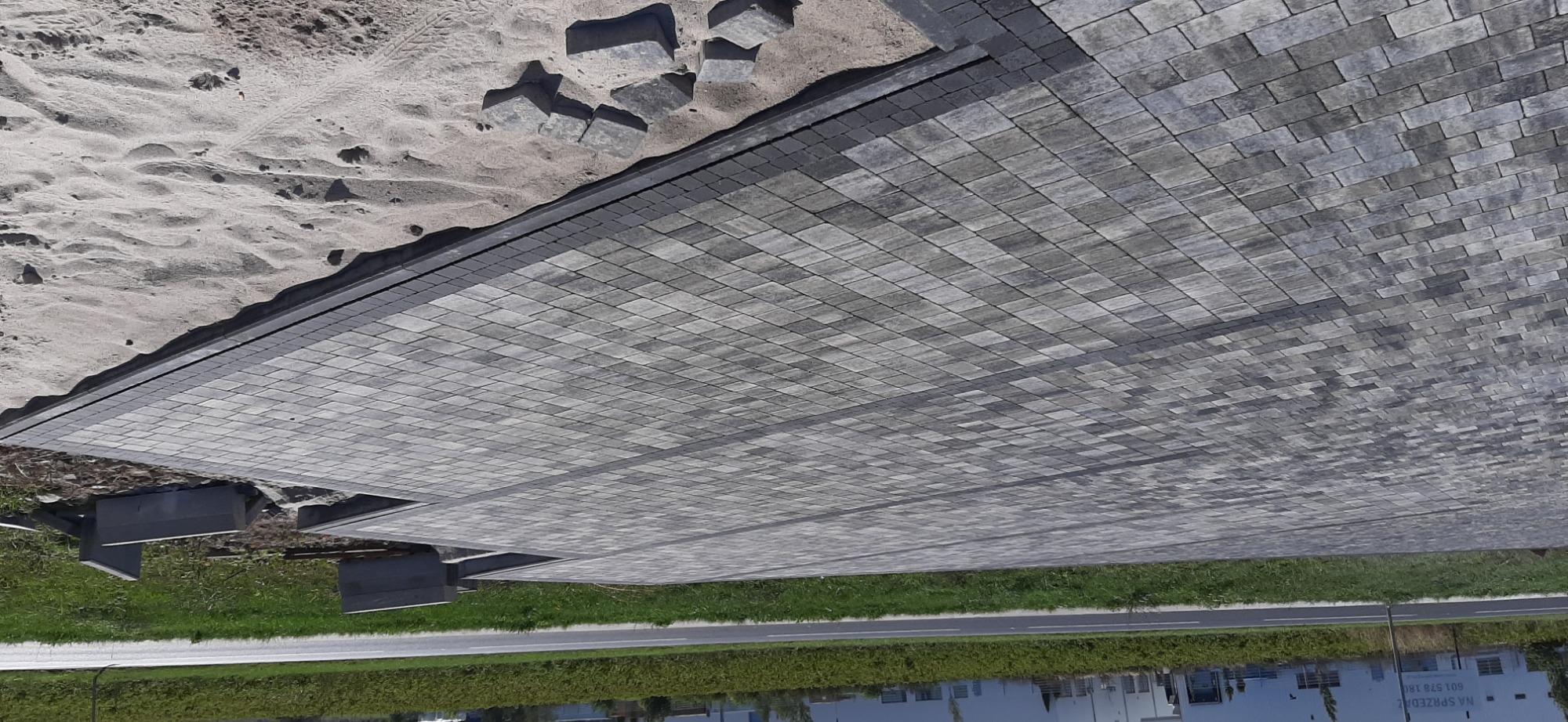 Ukończona nawierzchnia z szarej kostki brukowej ułożonej wzorem jodełki, z widocznymi niedokończonymi fragmentami i zapasem kostki na piasku.