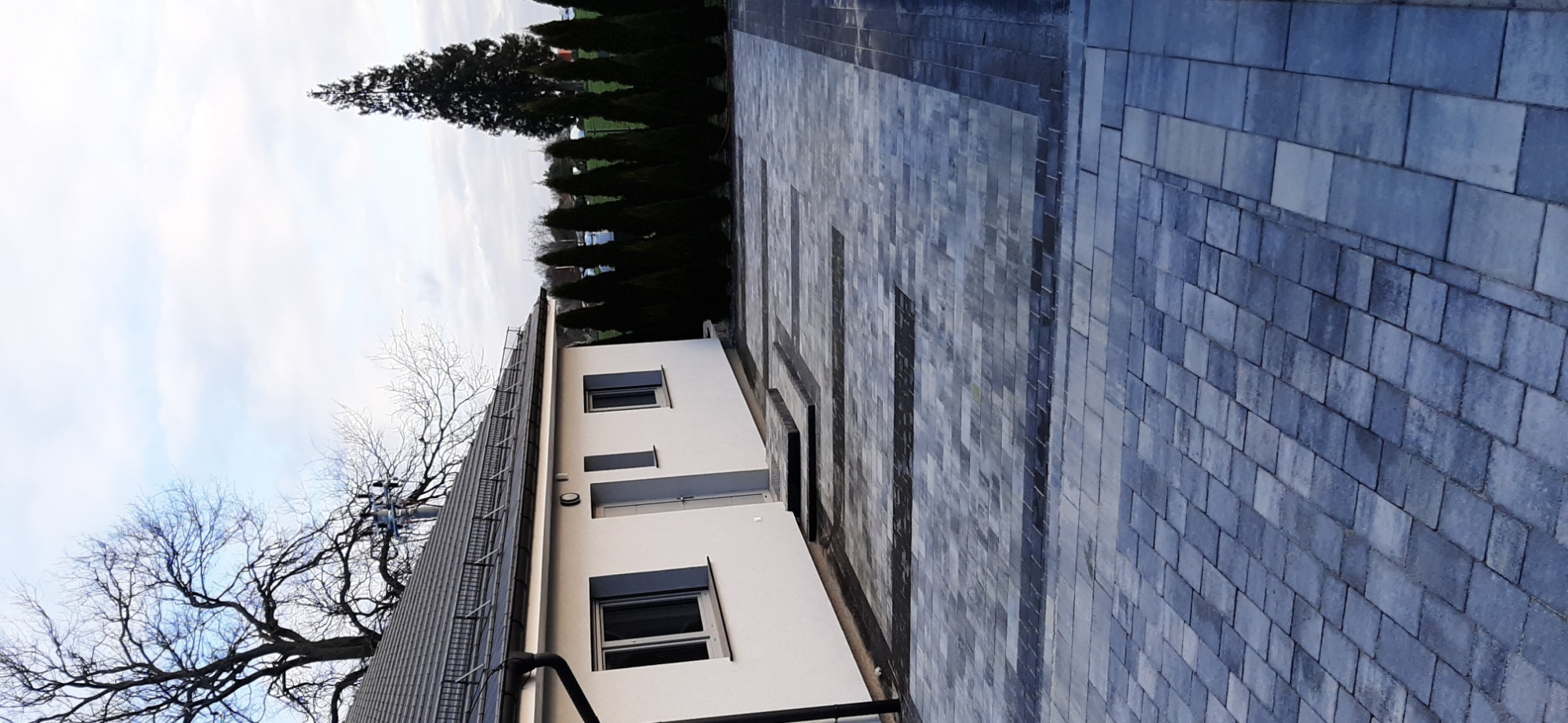 Ułożona szara kostka brukowa przed nowoczesnym, białym budynkiem z ciemnymi oknami i szarym dachem, widok z boku, w tle rząd tuj.